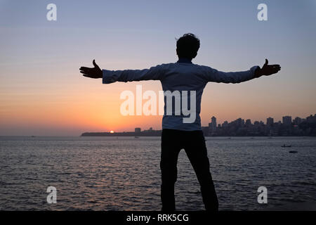 At Marine Drive by the Arabian Sea in Mumbai, India, a young Indian man poses with open arms in the direction of the setting sun, in a Bollywood pose Stock Photo