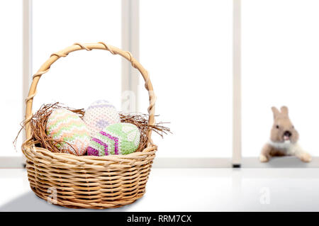 Colorful easter eggs on nest in wooden basket on the table and little bunny on window glass background. Happy Easter Stock Photo