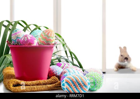 Colorful easter eggs on nest in the red bucket with fabric and table with palm branches and little bunny on window glass background. Happy Easter Stock Photo