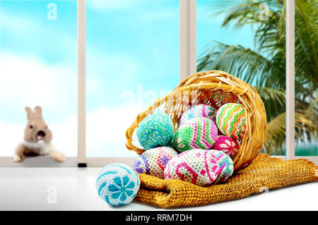 Colorful easter eggs spilled from wooden basket to fabric and table and little bunny on the window glass with palm tree and blue sky background. Happy Stock Photo