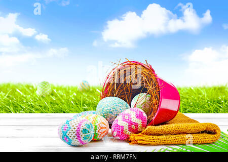 Colorful easter eggs spilled from nest on the red bucket to wooden table with fabric and palm leaf with hidden easter eggs on grass field on blue sky  Stock Photo