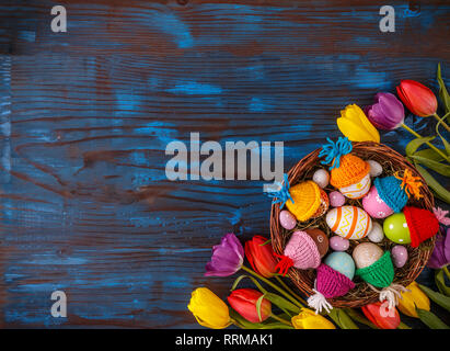 Easter composition with tulips and a basket of colored Easter eggs with little hats on blue wooden table Stock Photo