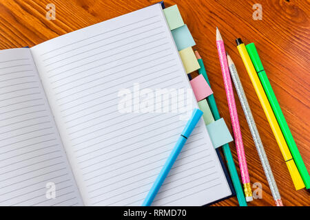 Opened notebook with marked pages and colorful pencils and felt-tip pens on wooden table. Stock Photo
