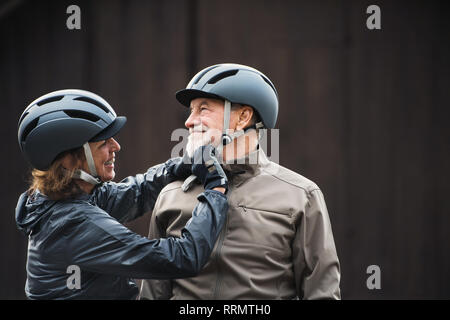 Happy active senior couple with bike helmets standing outdoors against dark background. Stock Photo