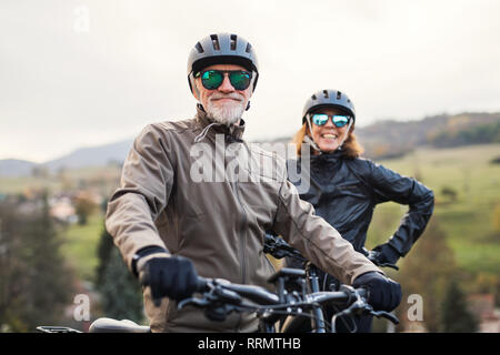 An active senior couple with helmets and electrobikes standing outdoors on a road in nature. Stock Photo