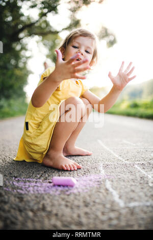 A small cute girl on a road in countryside in sunny summer nature, drawing with chalk. Stock Photo