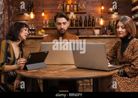 Smart man in casual style working on his laptop between two female colleagues in a coffee shop. Stock Photo