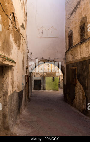 Dark morning narrow street with an arch city gate, made of stone bricks. White painted wall above the arch. Green part of the wall in the background.  Stock Photo