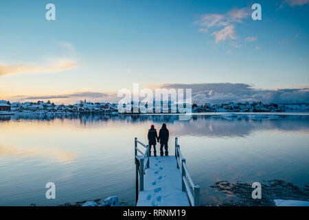 Couple holding hands at the edge of snowy dock overlooking waterfront village, Reine, Lofoten Islands, Norway Stock Photo