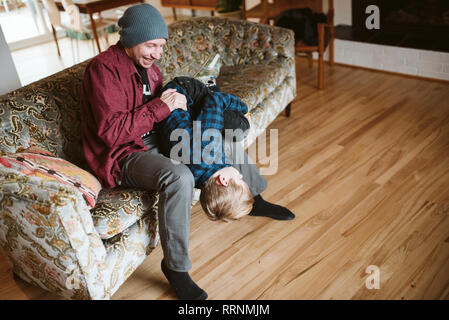 Playful father holding son upside-down on living room sofa Stock Photo