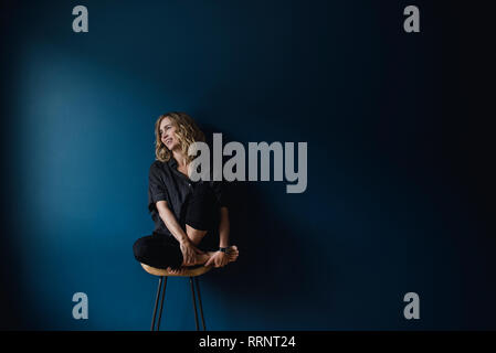 Portrait carefree woman sitting on stool against blue background Stock Photo