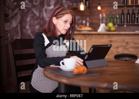 Beautiful smiling adult woman enjoying coffe while working on her tablet. Cozy vintage restaurant Stock Photo
