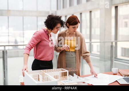 Two female young architects with model of a house standing in office, working and talking. Stock Photo