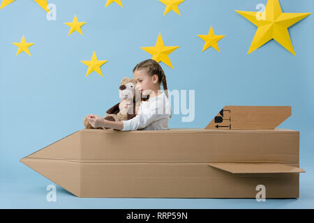 Kid holding teddy bear while playing with cardboard rocket on blue starry background Stock Photo