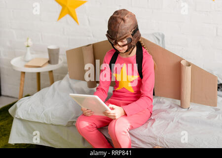Kid in pink clothes and flight helmet using digital tablet while sitting on bed Stock Photo