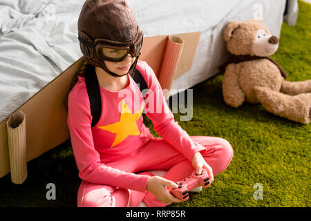 Child with cardboard wings sitting on carpet and playing video game with joystick Stock Photo