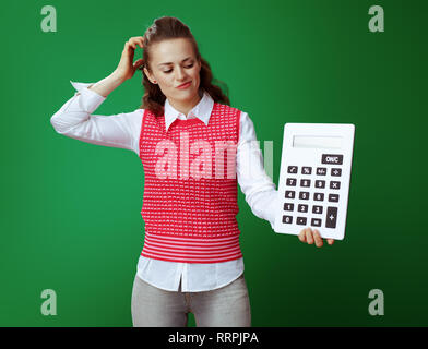 doubting healthy student in grey jeans and pink sleeveless shirt looking at big white calculator isolated on green. Financial challenges and expenditu Stock Photo