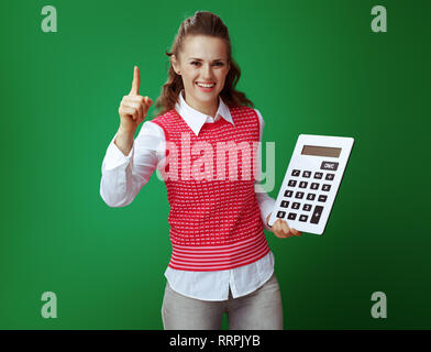 smiling healthy student in grey jeans and pink sleeveless shirt with big white calculator draws attention to something isolated on chalkboard green. F Stock Photo