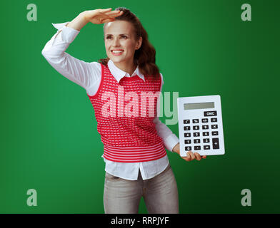 smiling fit learner woman in grey jeans and pink sleeveless shirt with big white calculator looking into the distance isolated on green background. Fi Stock Photo