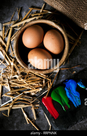 high angle view of some brown chicken eggs in a boxwood bowl, some paint of different colors on a black chalk palette, and some straw stalks on a gray Stock Photo