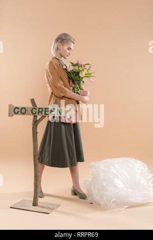 blonde girl in eco clothing holding vase with flowers near sign with go green lettering on beige background, environmental saving concept Stock Photo