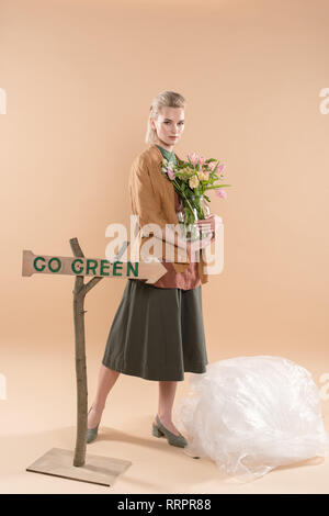 attractive girl in eco clothing holding vase with flowers near sign with go green lettering on beige background, environmental saving concept Stock Photo