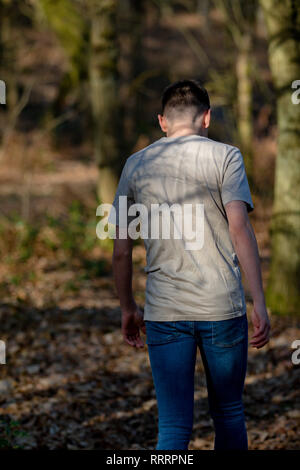 Teenage caucasian boy walking away through woodland on a warm spring day looking over his shoulder Stock Photo