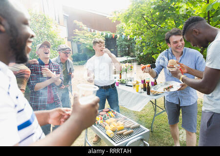 Male friends laughing and eating around barbecue grill in backyard Stock Photo