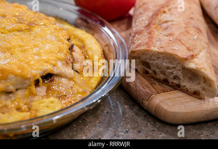 Baking in a glass tray in electrical oven a layered meat pie with mushrooms and vegetables, covered with melted cheese ready to eat and long loaf on c Stock Photo