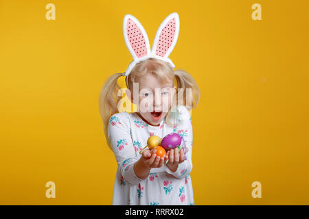 Cute adorable Caucasian blonde girl in white dress with pink  Easter bunny ears holding eggs in studio on yellow background. Funny surprised kid child Stock Photo