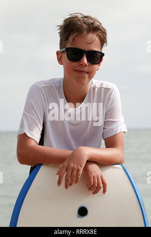 A young surfer boy poses for a portrait after hitting the waves while wearing sunglasses and leaning on a boogie board. Stock Photo