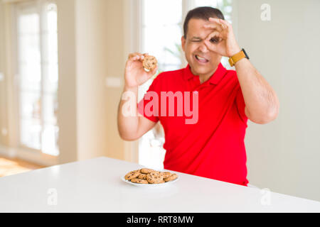 Middle age man eating chocolate chips cookies at home with happy face smiling doing ok sign with hand on eye looking through fingers Stock Photo
