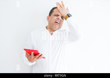 Middle age man using digital table over white wall stressed with hand on head, shocked with shame and surprise face, angry and frustrated. Fear and up Stock Photo