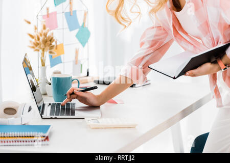 Partial view of woman with notebook and pen typing on laptop keyboard Stock Photo