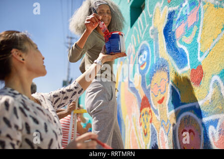 Female volunteers painting vibrant community mural on sunny urban wall Stock Photo