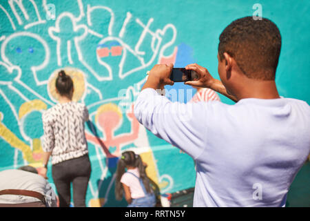 Man with camera phone photographing community mural on sunny wall Stock Photo