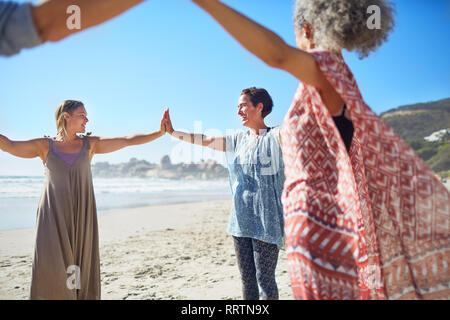Women joining hands in circle on sunny beach during yoga retreat Stock Photo