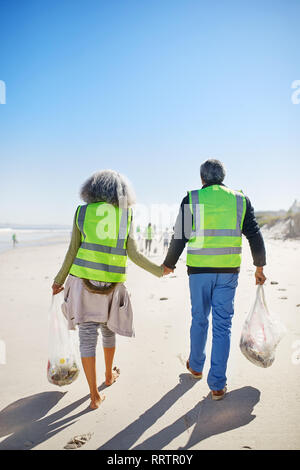 Affectionate senior couple volunteers cleaning up litter on sunny, wet sand beach Stock Photo