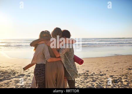 Female friends with yoga mats hugging on sunny beach Stock Photo