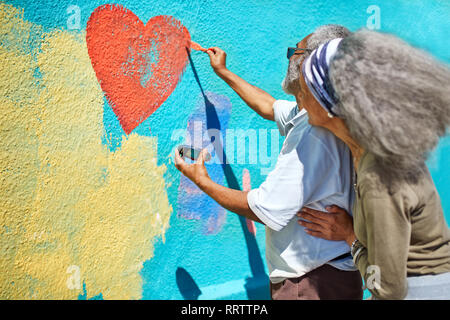 Senior couple painting heart-shape mural on sunny wall Stock Photo