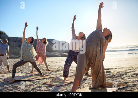 Group practicing yoga reverse warrior pose on sunny beach during yoga retreat Stock Photo