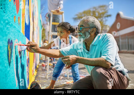 Grandfather and granddaughter volunteers painting vibrant mural on sunny urban wall Stock Photo