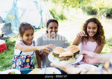 Happy mother and daughters reaching for barbecue hamburgers at campsite Stock Photo