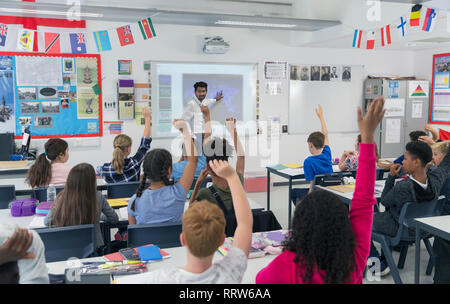 Male teacher leading lesson at projection screen in classroom with students raising hands Stock Photo