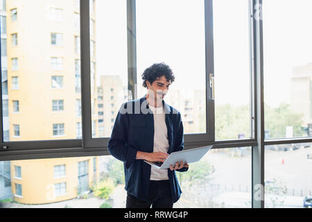Man standing, using laptop at urban apartment window Stock Photo