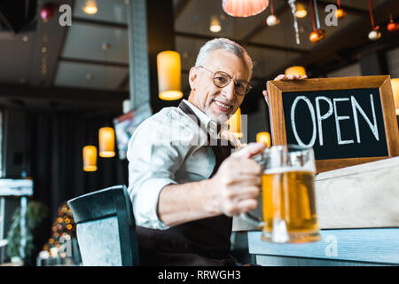 smiling senior owner of pub holding open sign and glass of beer Stock Photo