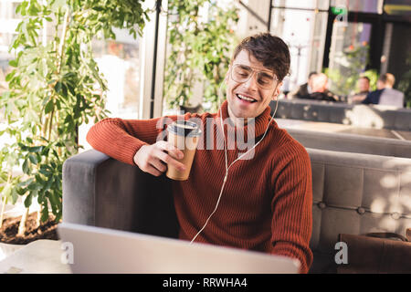 smiling man listening music in earphones while using laptop and holding disposable cup in cafe Stock Photo