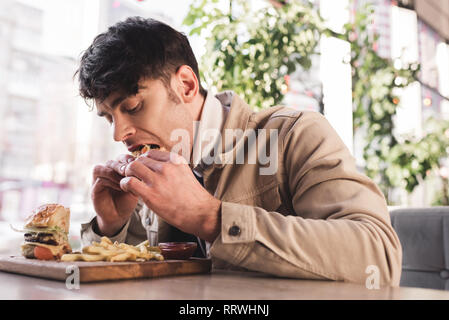 selective focus of young man eating tasty hamburger near french fries on cutting board in cafe Stock Photo