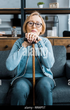 upset senior woman in glasses sitting on couch, looking at camera and leaning on walking stick in living room Stock Photo