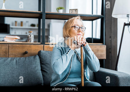 upset senior woman in glasses sitting on couch and leaning on walking stick in living room Stock Photo